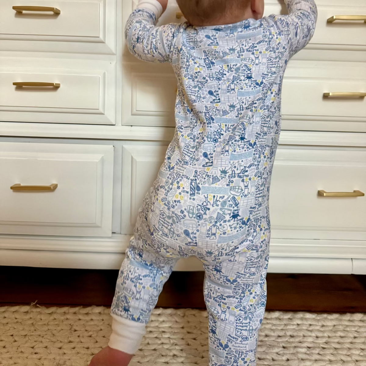 Rear view of tennis zip baby onesie showcasing a blue hand-drawn tennis print with white cabinetry in the background