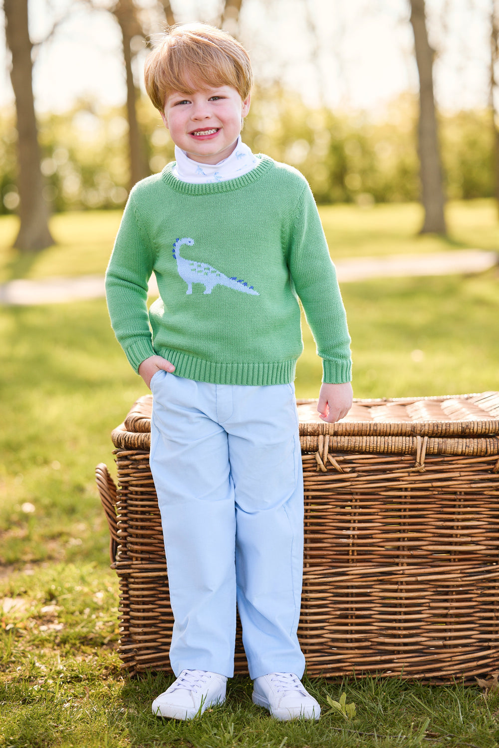 Boy standing outdoors wearing a green sweater with blue dinosaur, white turtleneck, and light blue corduroy pants