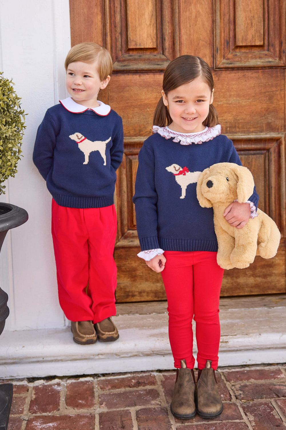 Front view of girl’s navy Intarsia sweater with cream Labrador and red bow collar, paired with red leggings at wooden door