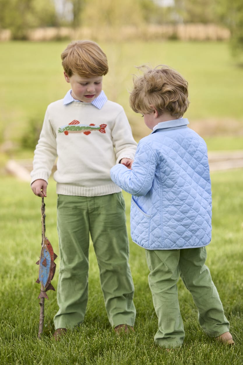 Little English traditional children's clothing.  Boys cream crew neck sweater with fish motif layered over light blue striped polo and paired with watercress green corduroy pants. Little boy wearing light blue quilted jacket with watercress green corduroy pull on pants