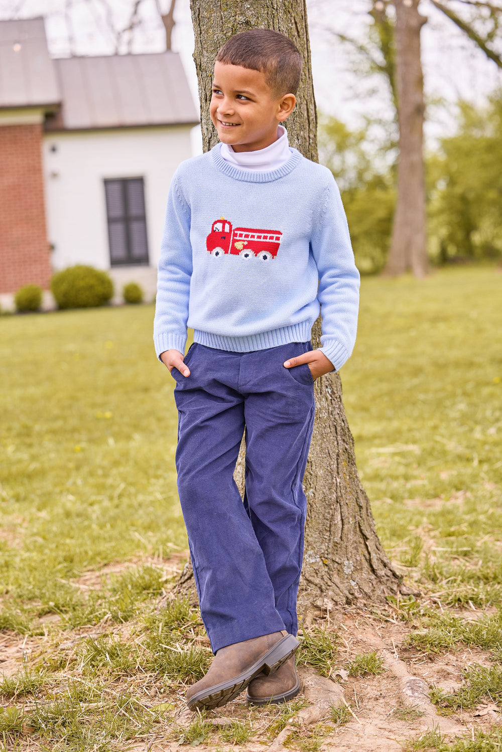 Boy leaning against a tree wearing a light blue fire truck intarsia sweater over a white turtleneck and navy corduroy pants
