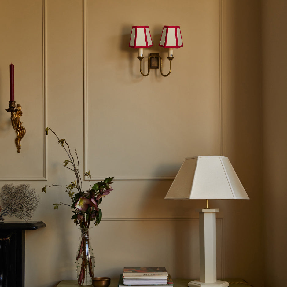 Hexagon linen candle shades with red trim on wall sconces above a table lamp with off-white hexagon shade and floral vase in a neutral room