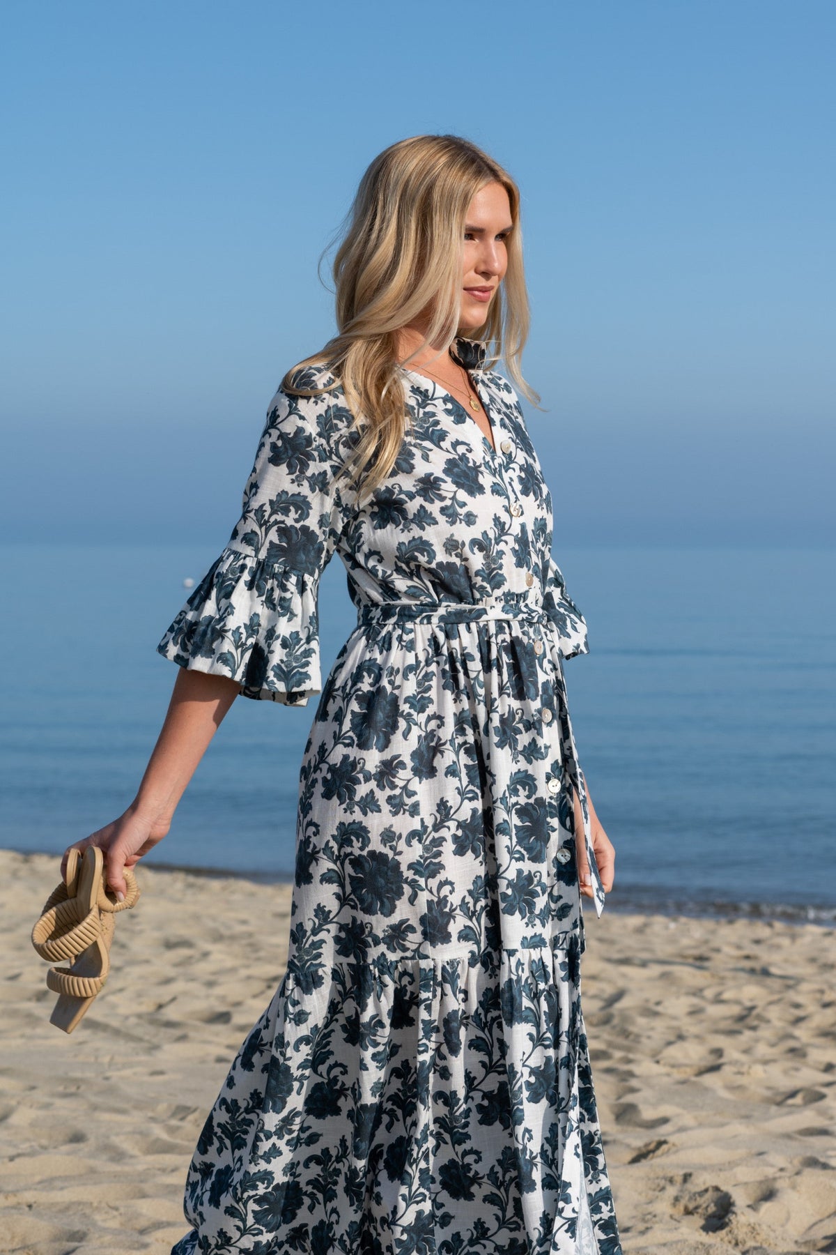 Three-quarter view of Joanna Dress in Blue Dahlia on a model showcasing bell sleeves, flounced skirt, button-front bodice and removable sash on a sandy beach