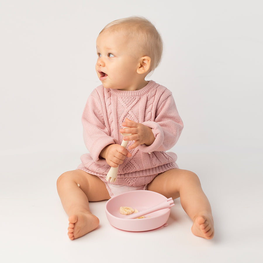 Baby sitting on a white floor holding a spoon, looking to the left with a pink silicone suction bowl and fork nearby