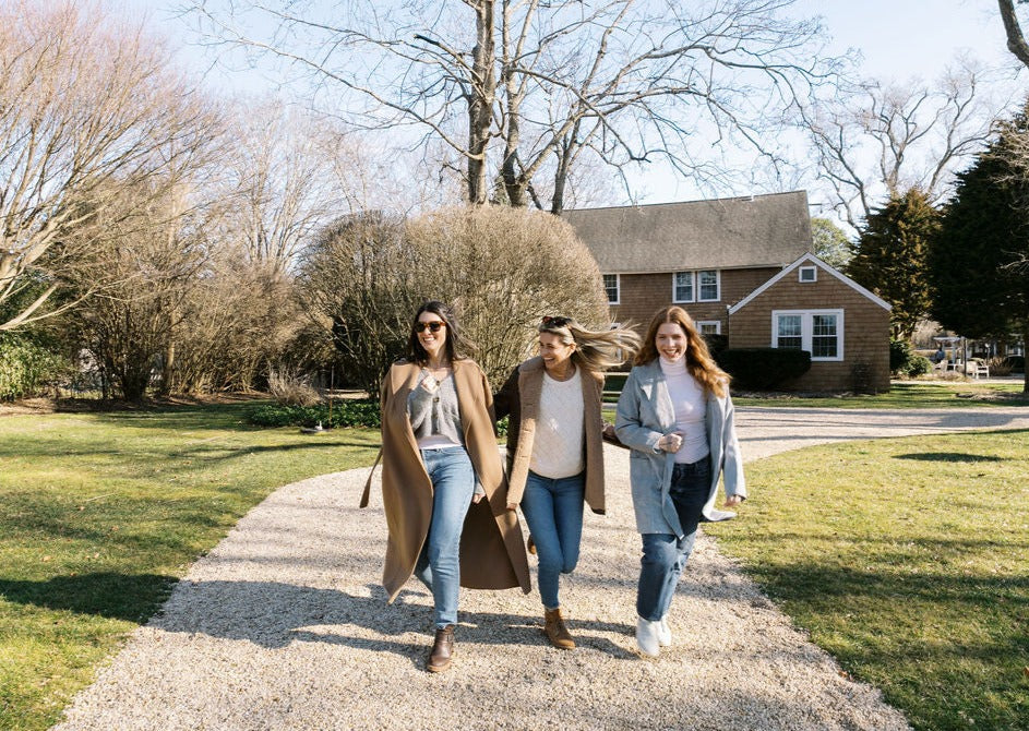 Three women walking arm-in-arm along a gravel driveway in front of a Hamptons house on a sunny day