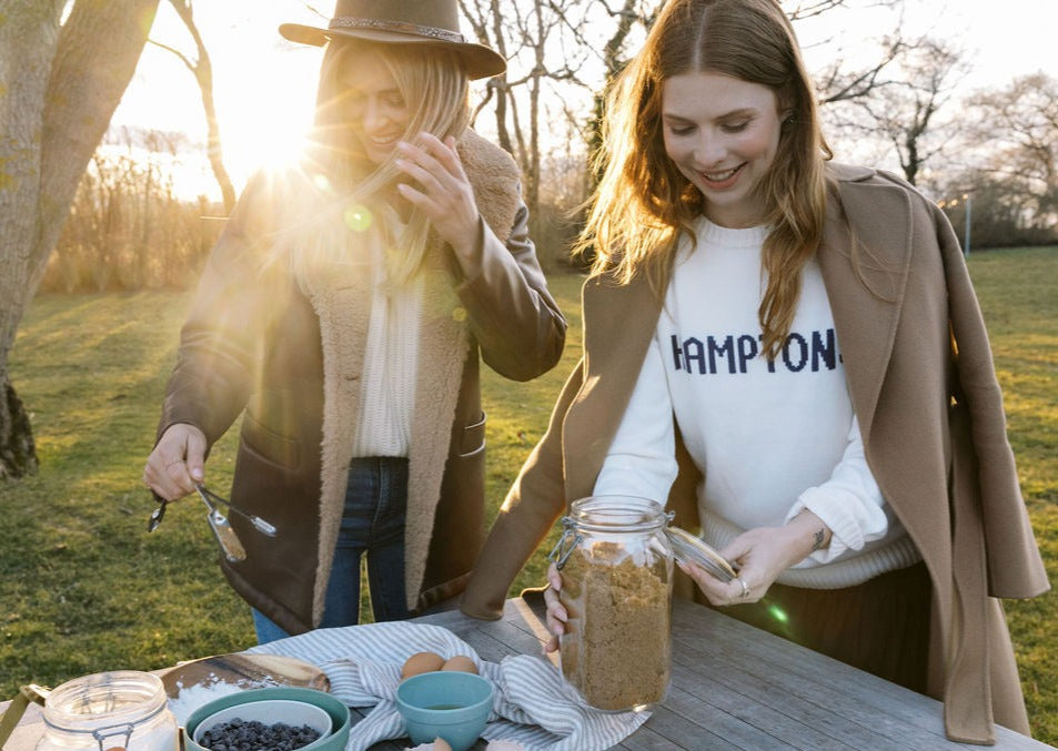 Two women arranging picnic bowls and a jar at a wooden table in a sunlit field, one wearing a white sweater that reads HAMPTONS