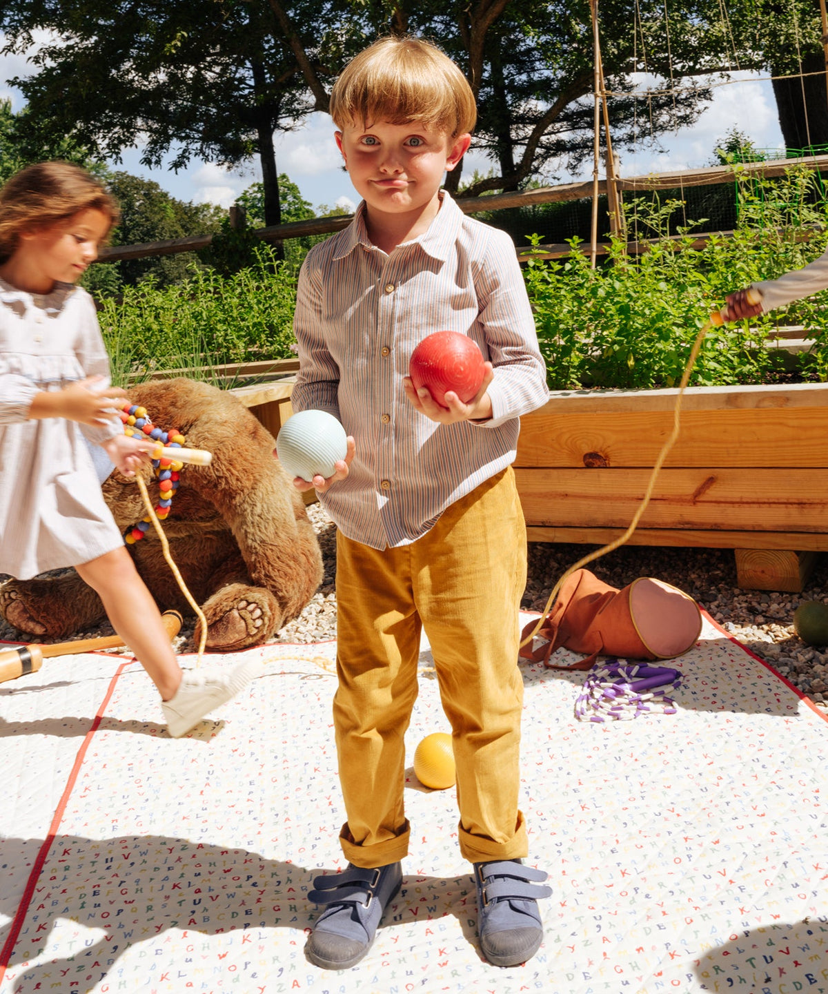 Child holding red and pale blue balls on a picnic blanket in a garden, wearing Jeffie shirt in indigo linen stripe