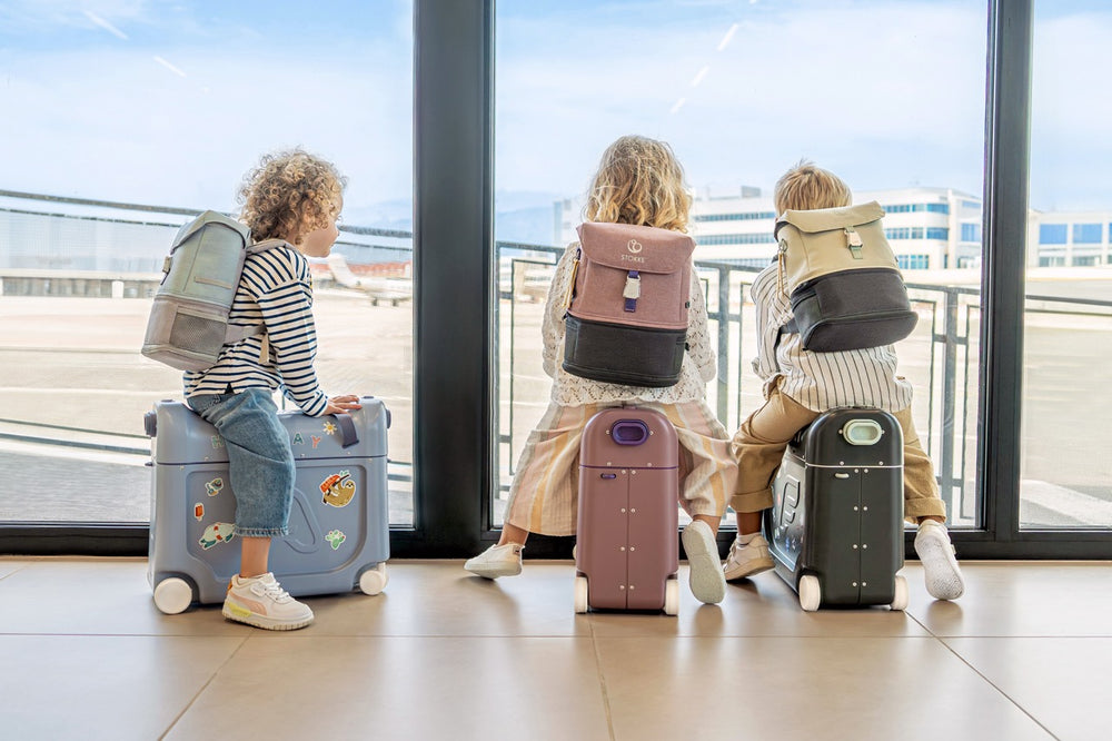 Three children sitting on JetKids ride-on suitcases at an airport window, each wearing a backpack