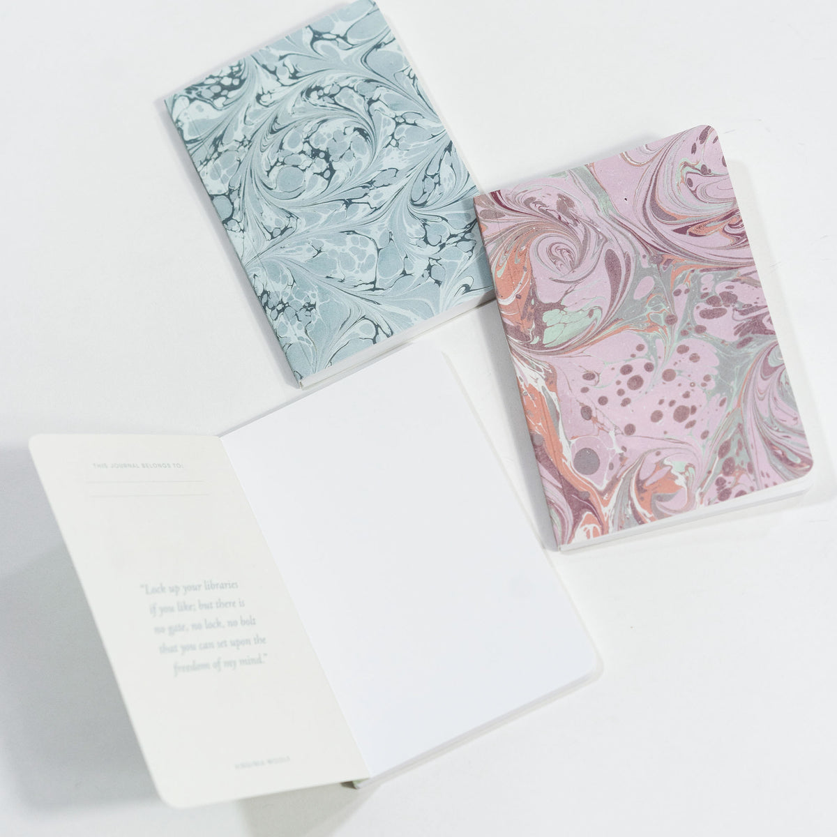 Top-down view of three marble pocket journals showing blue and pink marbled covers and one open notebook on white background