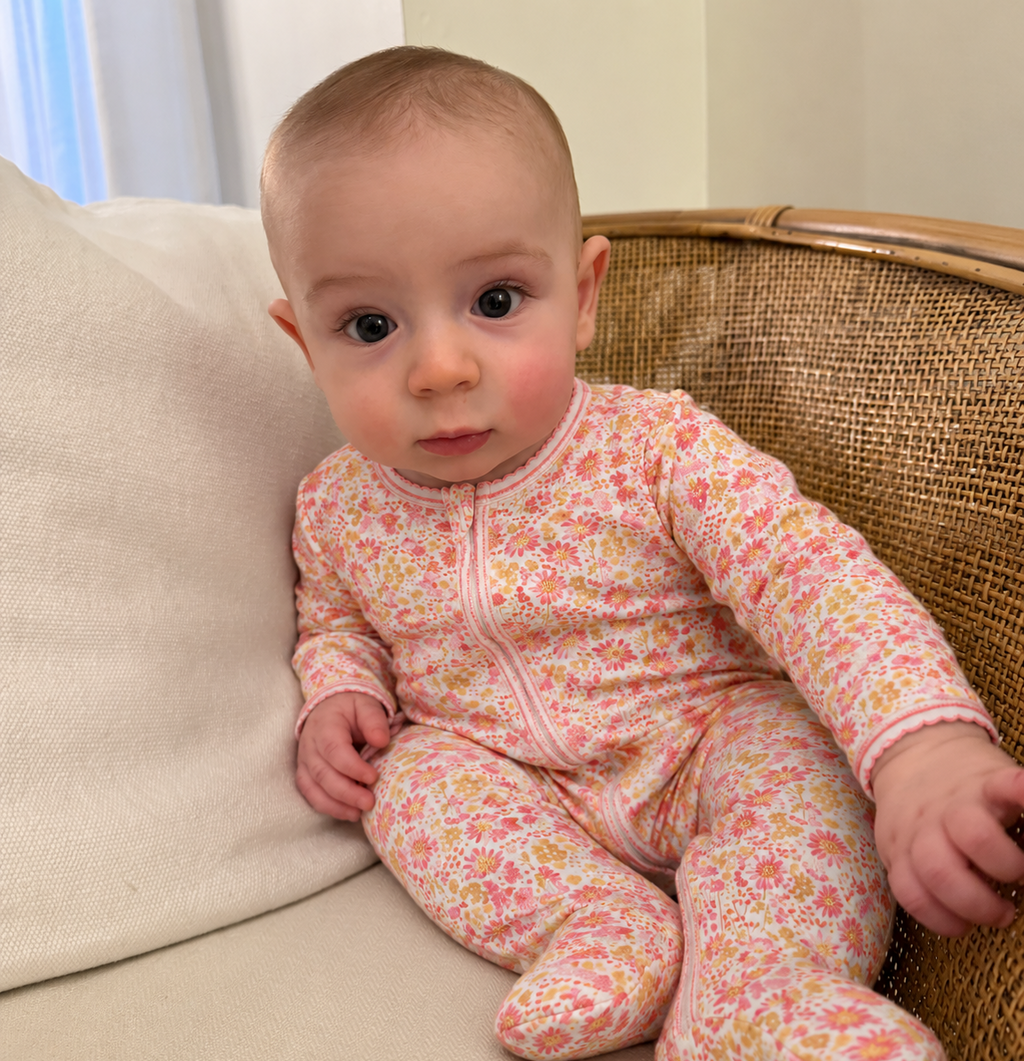 Front view of pink floral baby onesie showcasing long sleeves and footed legs on a seated baby against a cream chair