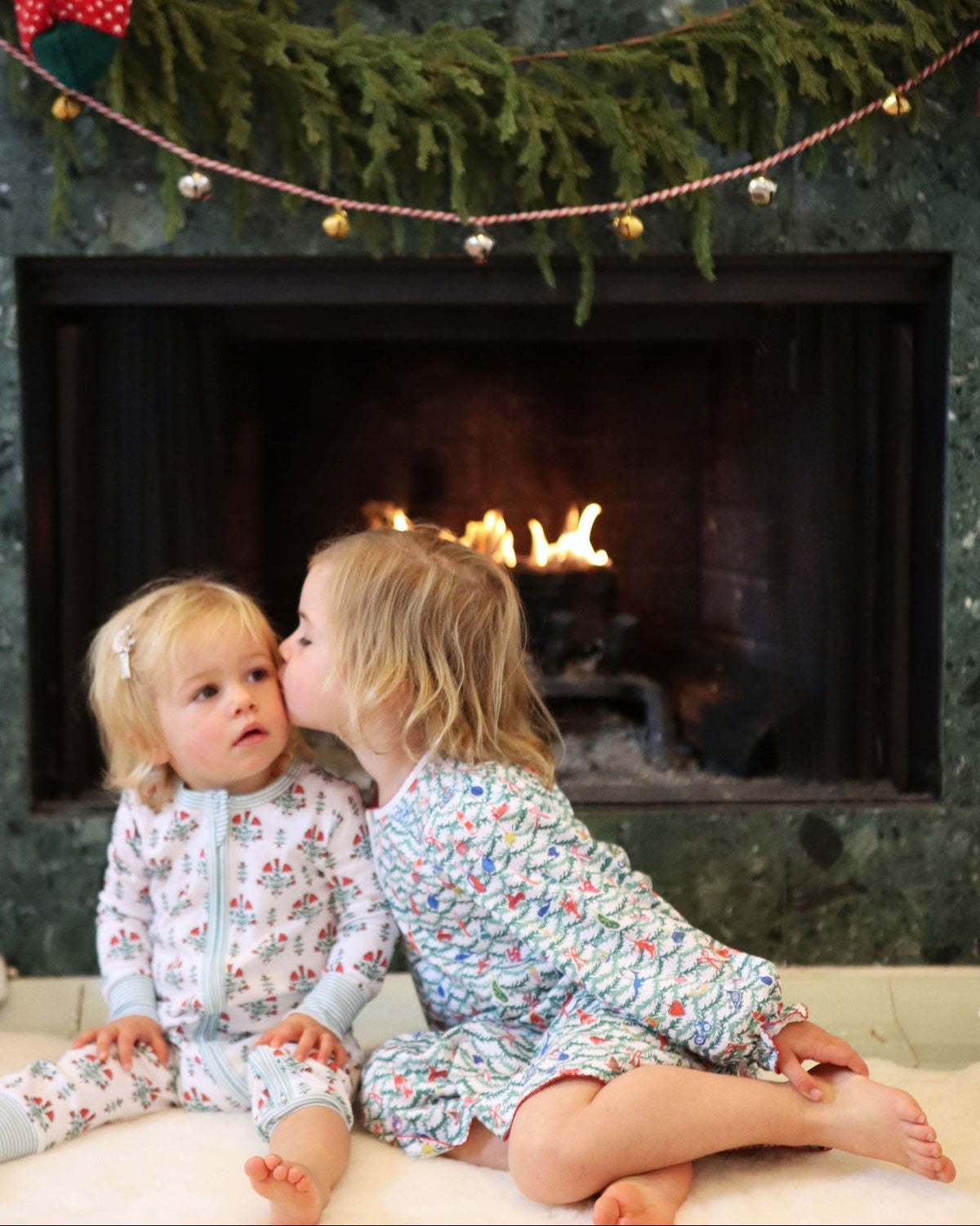 Two girls in Christmas-print lounge dresses sit beside a lit fireplace
