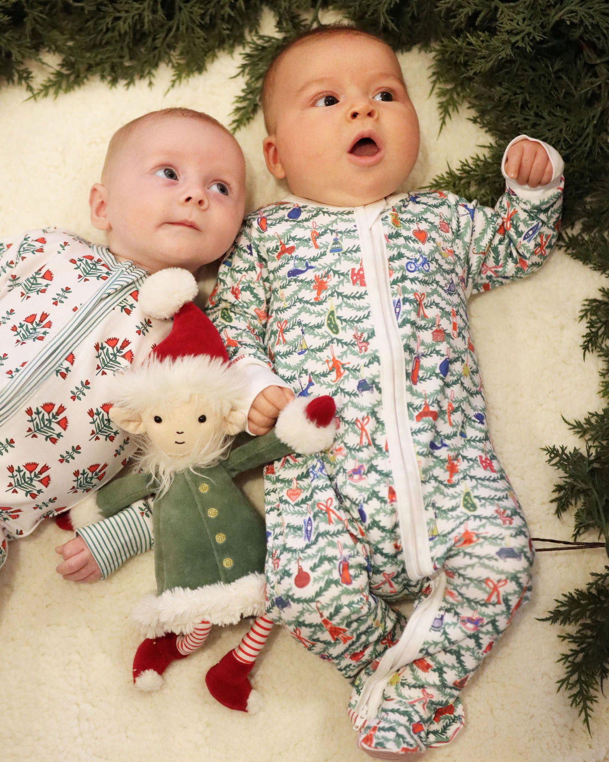 Two babies wear Christmas tree print zip onesies beneath evergreen branches beside a holiday teddy bear