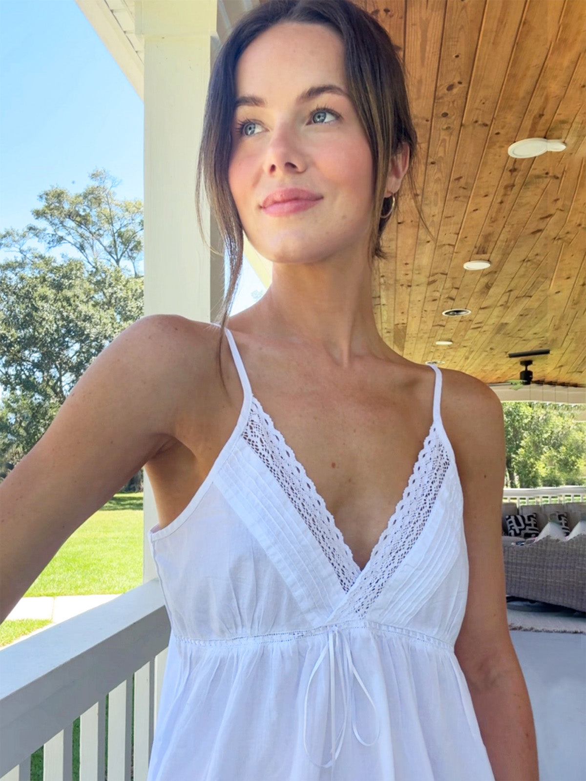 Front view of Julie white cotton nightgown showcasing V-neck lace trim, adjustable spaghetti straps and ribbon tie on a porch