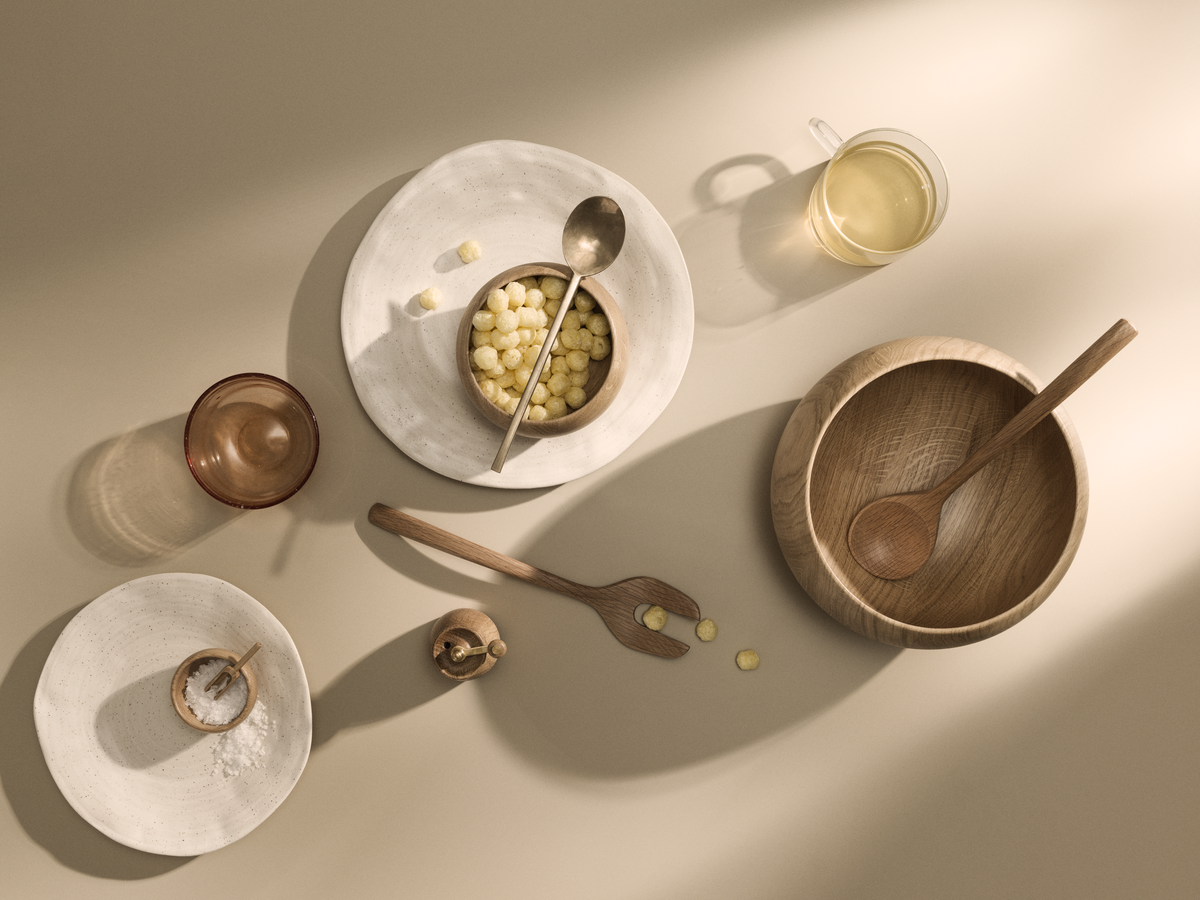 Overhead view of Kay Bojesen oak serving bowl with wooden spoon, on beige surface with plates and glass