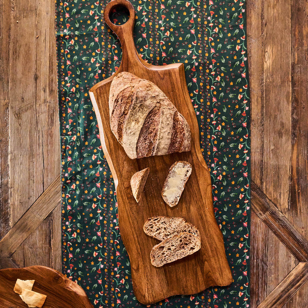 Top-down view of Puro long serving board in teak showcasing hand-carved grain and elongated organic shape on a green floral cloth
