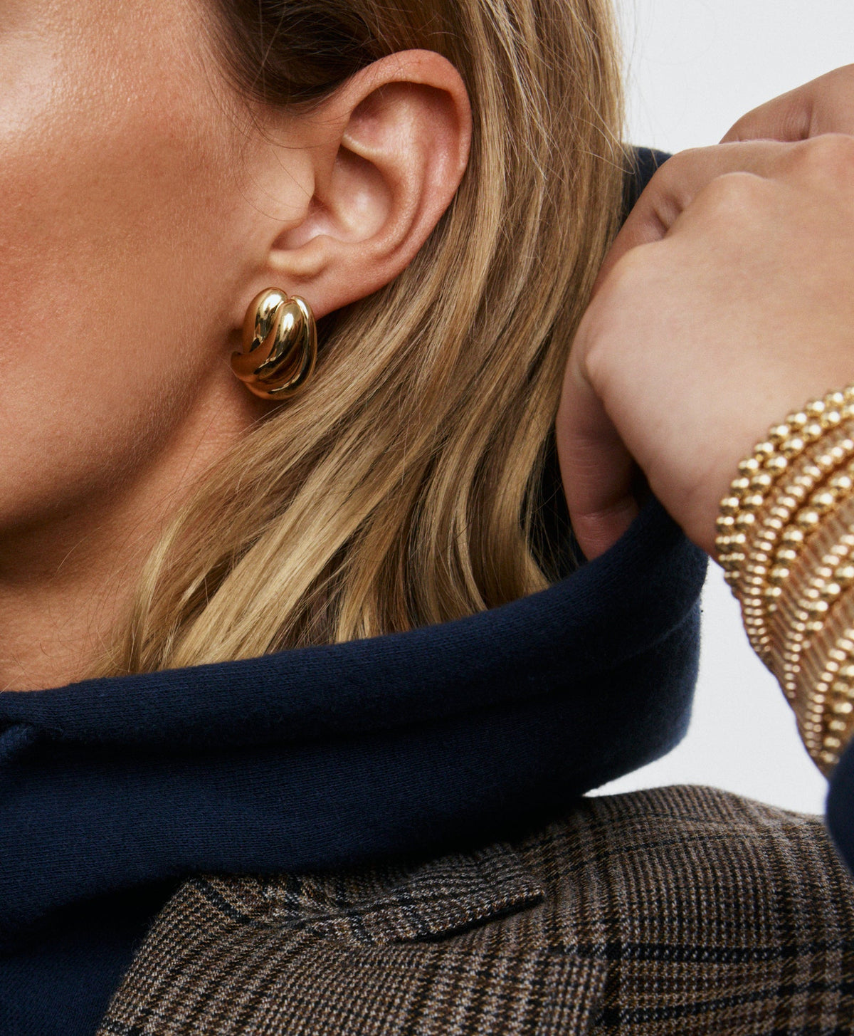Close-up side view of Knot Earrings on model, gold-tone twisted knot post earrings worn with navy hoodie and plaid jacket