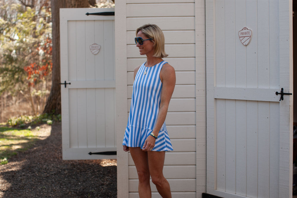 Side view of All-Sport Buffy Dress on model showcasing blue-and-white vertical stripes, sleeveless silhouette and flared hem against a white garden shed door