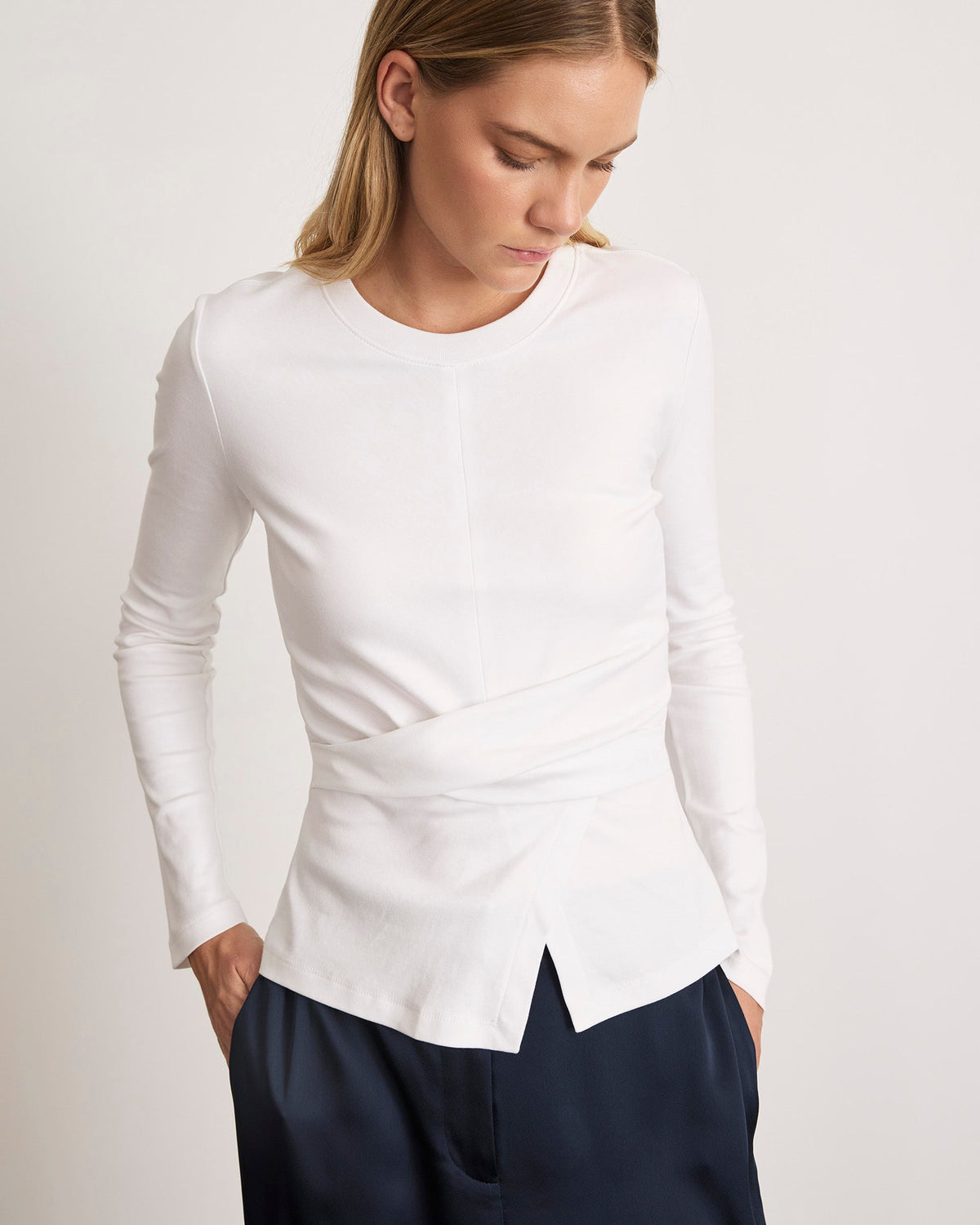 Three-quarter view of Long Sleeve Kelsey top in optic white with twist waist detail, crew neckline, long sleeves, and front hem slit on neutral background