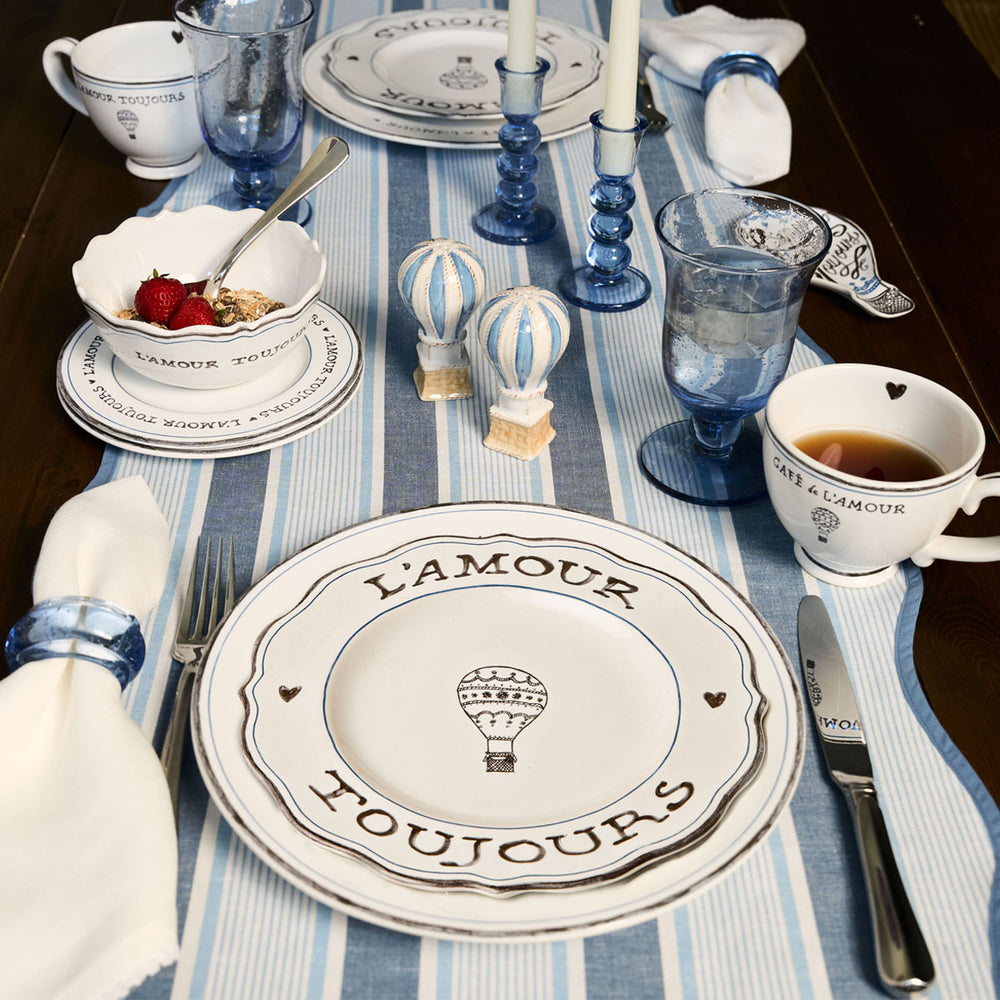 Top-down view of L’Amour Toujours 16-piece place setting showcasing hand-sketched hot air balloons and heart motifs on white stoneware with a blue striped table runner