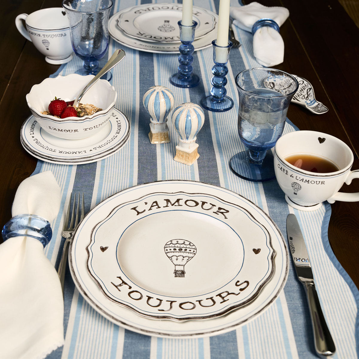 Top-down view of L’Amour Toujours 16-piece place setting showcasing hand-sketched hot air balloons and heart motifs on white stoneware with a blue striped table runner