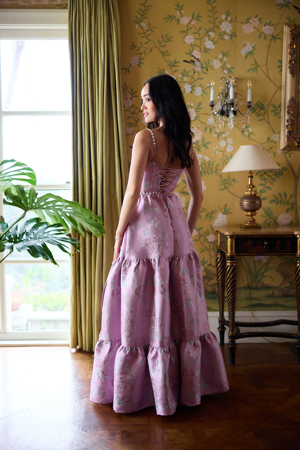 Back view of The Lucetta Dress in Orchid Baroque Floral showcasing lace-up corset and tiered skirt in an elegant room