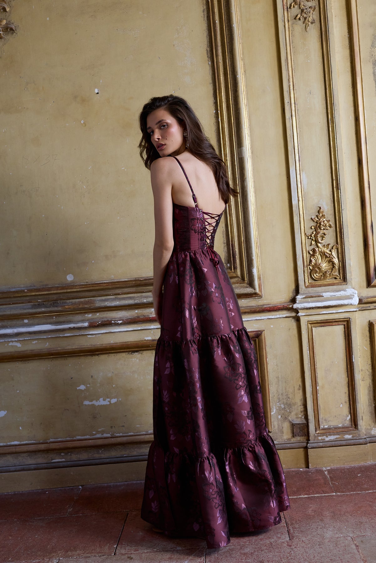Back view of The Lucetta Dress in Plum Baroque Floral showcasing lace-up corset and tiered skirt against an ornate wall