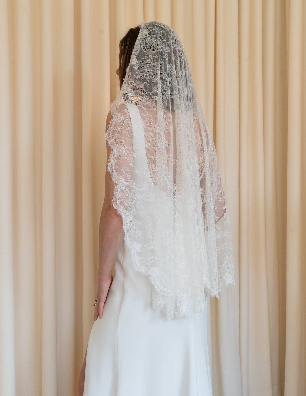 Rear view of Lace Mantilla veil on model showcasing scalloped Chantilly lace edge and fingertip length against cream curtains