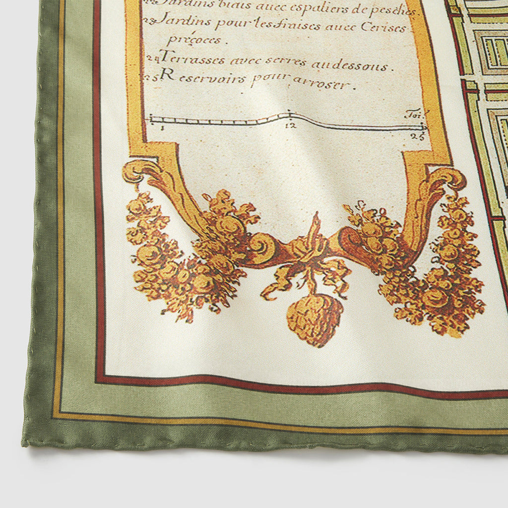 A close-up, detailed photograph of the corner of a silk scarf. It features an antique-style illustration on a cream background, showing a decorative golden-brown cartouche filled with fruit, grain, and leaves. Above the illustration, elegant French script reads "Jardins pour les fraises avec Cerises..." and "Terrasses avec serres au-dessous." The scarf is finished with a hand-rolled, solid sage green border.