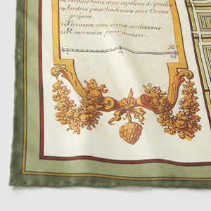 A close-up, detailed photograph of the corner of a silk scarf. It features an antique-style illustration on a cream background, showing a decorative golden-brown cartouche filled with fruit, grain, and leaves. Above the illustration, elegant French script reads "Jardins pour les fraises avec Cerises..." and "Terrasses avec serres au-dessous." The scarf is finished with a hand-rolled, solid sage green border.