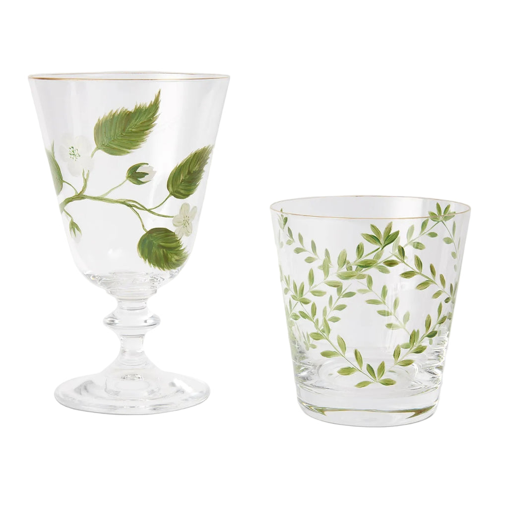 A product photograph displaying two clear glassware pieces side by side against a light gray background, both featuring gold rims and delicate hand-painted botanical designs. On the left is an elegant, stemmed footed glass decorated with a branch of green leaves, white blossoms, and a single green apple. On the right is a matching tapered tumbler adorned with a symmetrical wreath-like pattern of slender green branches and fine leaves.