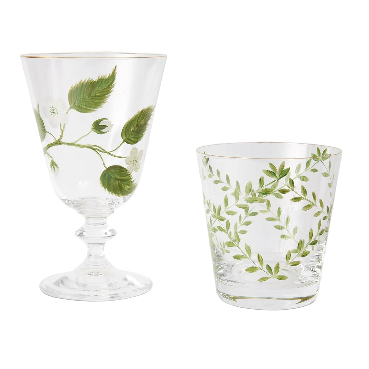 A product photograph displaying two clear glassware pieces side by side against a light gray background, both featuring gold rims and delicate hand-painted botanical designs. On the left is an elegant, stemmed footed glass decorated with a branch of green leaves, white blossoms, and a single green apple. On the right is a matching tapered tumbler adorned with a symmetrical wreath-like pattern of slender green branches and fine leaves.