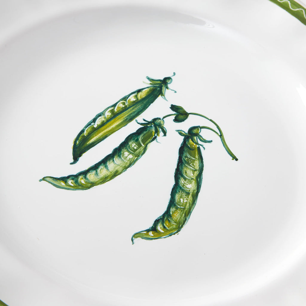 A close-up product photograph highlighting the center of a white ceramic plate. The image features a detailed, hand-painted botanical illustration of three vibrant green pea pods clustered together on a vine, with one pod partially open to reveal the small peas inside. A thin, wavy green zigzag line is visible along the top edge of the plate's rim.