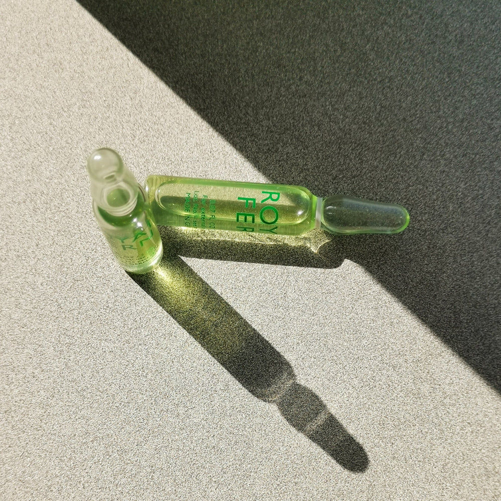 Top-down view of two Phytoactive Illuminating Ampoules showing clear liquid with one ampoule opened and a strong diagonal shadow on a textured gray surface