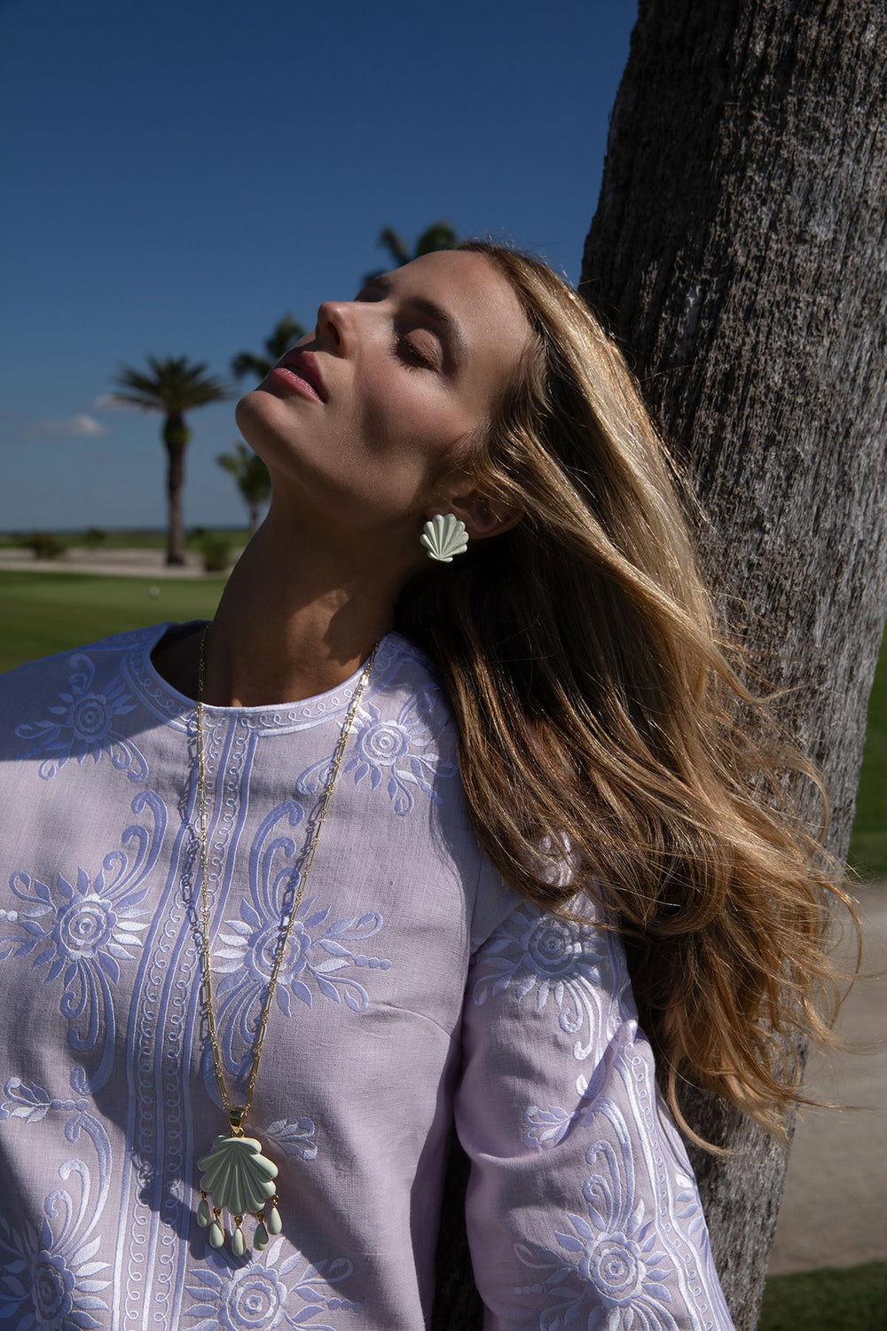 Close-up of Sirena Stud clip-on ivory scallop shell earring worn on model with palm tree and blue sky background