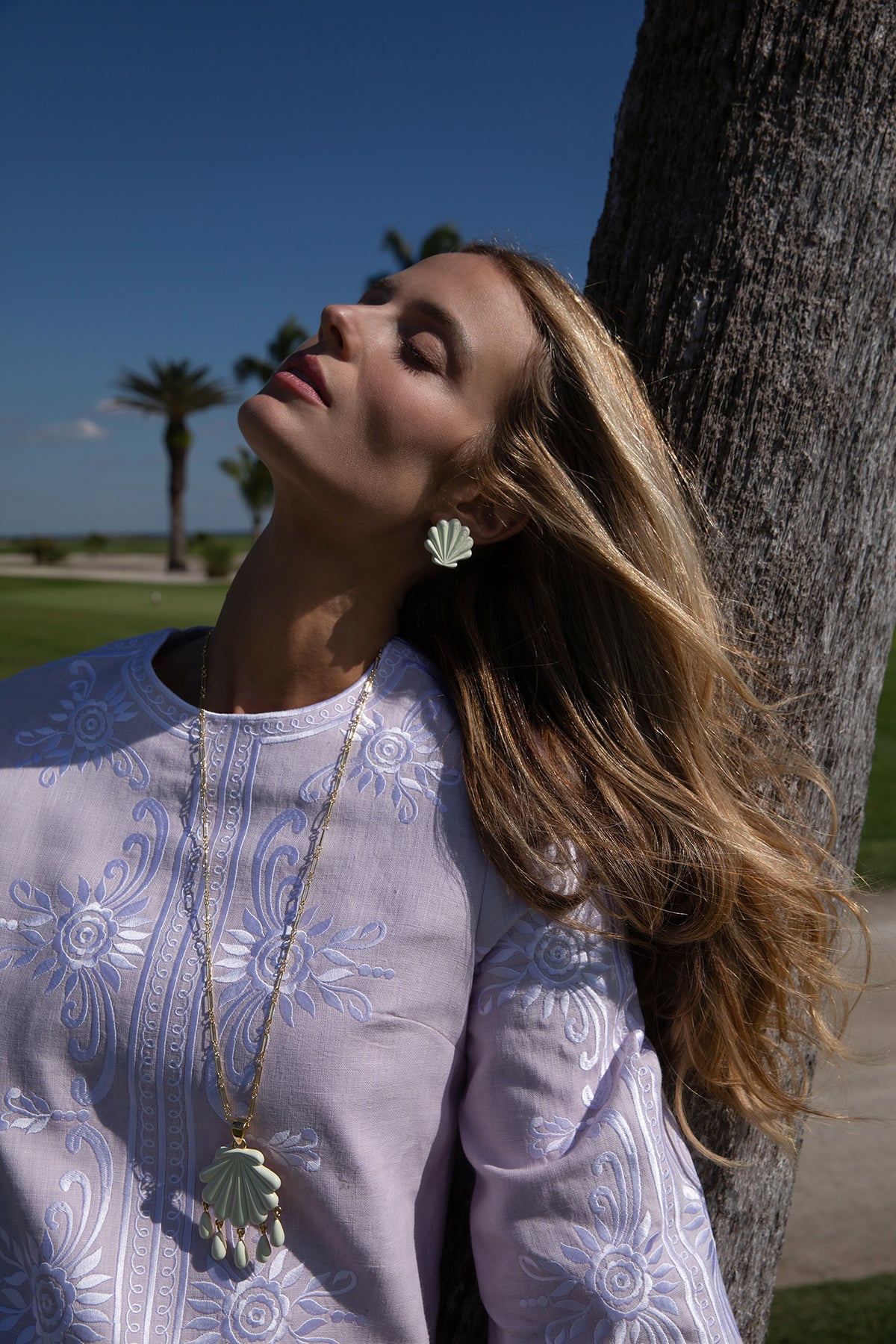 Close-up of Sirena Stud clip-on ivory scallop shell earring worn on model with palm tree and blue sky background