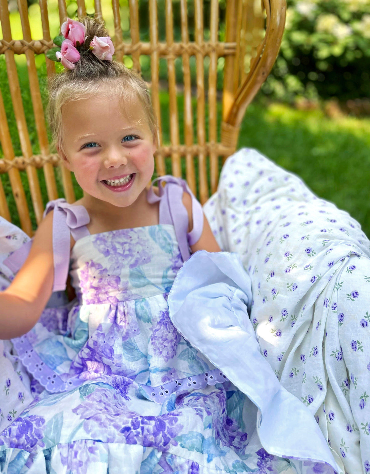 Child wearing the Blooming Lavender Hydrangea Dress seated in a wicker chair outdoors, showing floral print and shoulder bows