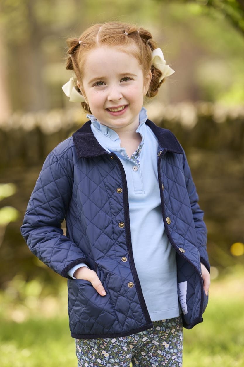 Little English traditional children's clothing.  Long sleeve Hastings polo in light blue with ruffle collar and green floral detailing. Paired with green floral leggings for little girls with navy quilted jacket layered over