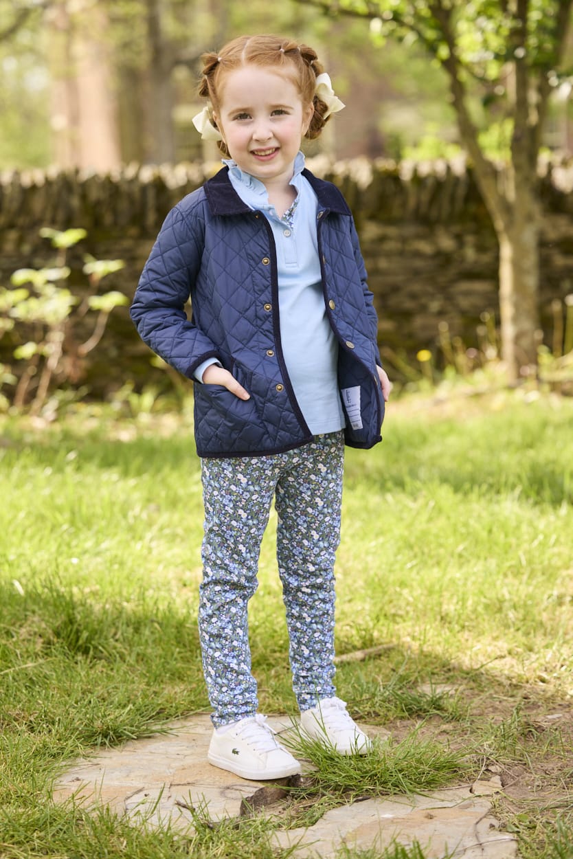 Little English traditional children's clothing.  Long sleeve Hastings polo in light blue with ruffle collar and green floral detailing. Paired with green floral leggings for little girls with navy quilted jacket layered over