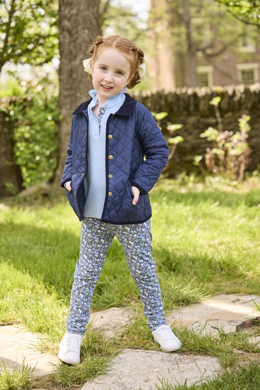 Little English traditional children's clothing.  Long sleeve Hastings polo in light blue with ruffle collar and green floral detailing. Paired with green floral leggings for little girls with navy quilted jacket layered over