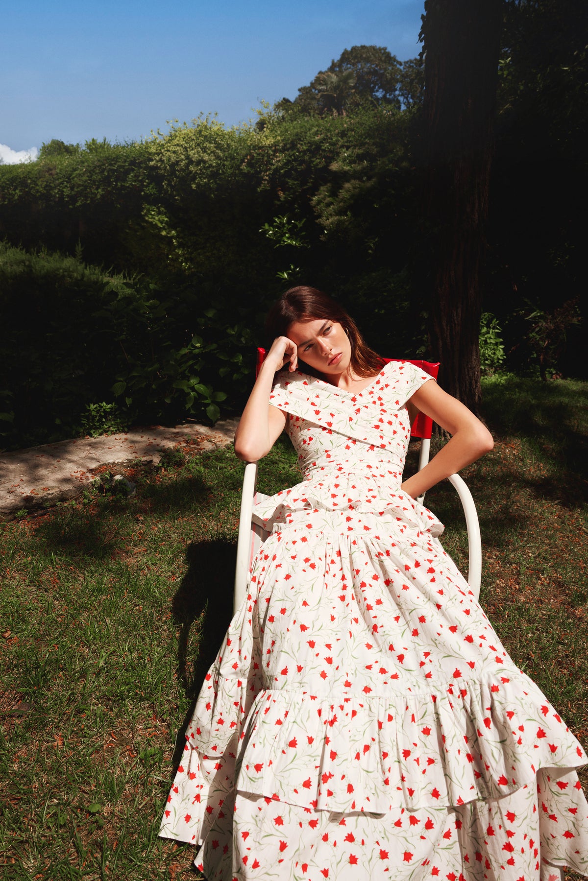 Model relaxing in a garden wearing a white floral outfit beside a red chair