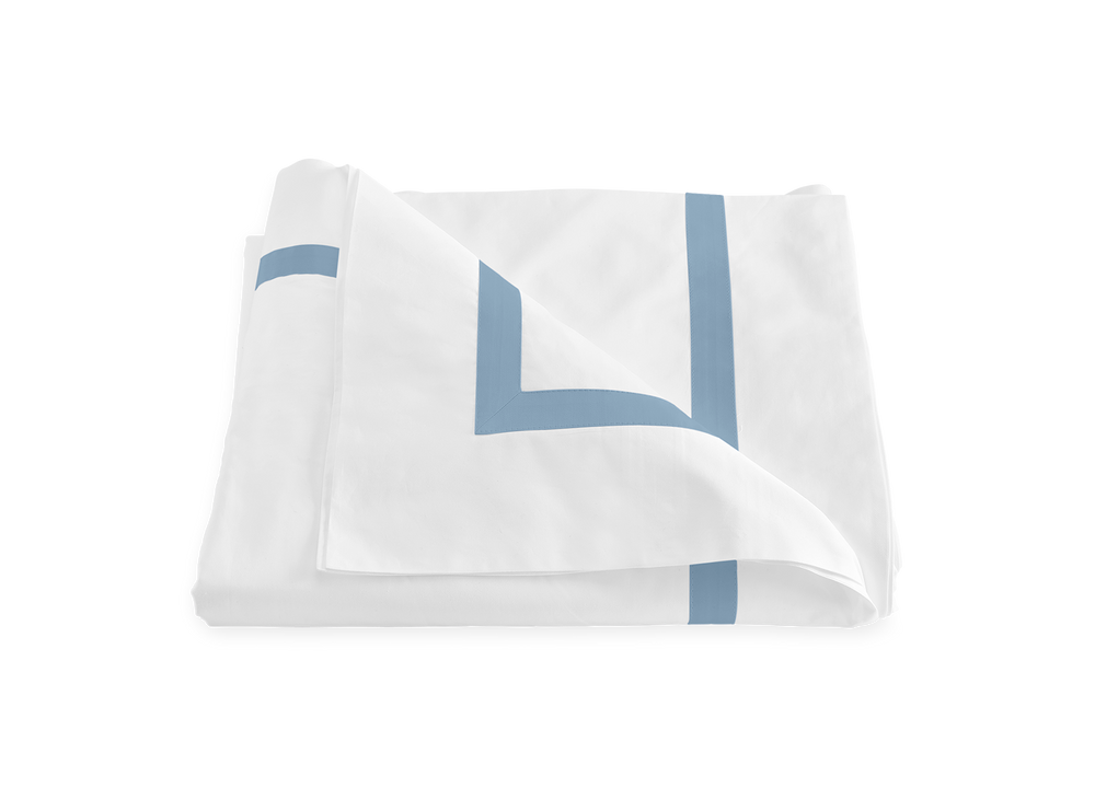 Three-quarter view of folded Lowell duvet cover in hazy blue showing double border trim on white fabric