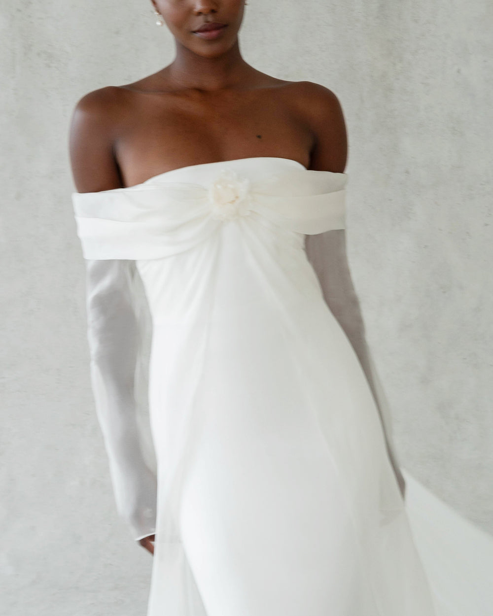 Front view of Ludlow Flower silk organza rosette clipped to an off-the-shoulder white gown with sheer long sleeves against a light gray backdrop