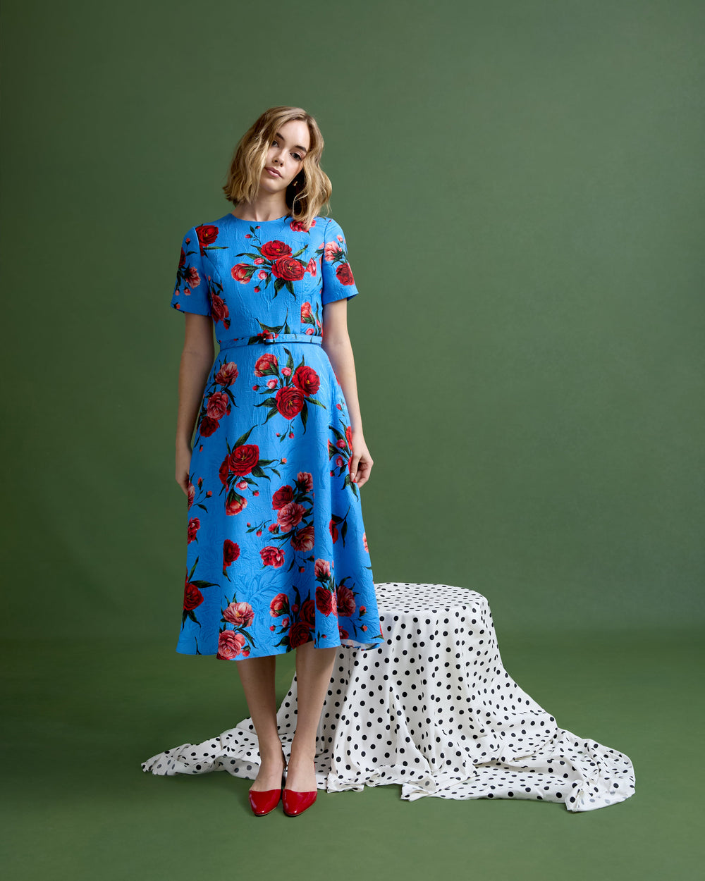 Front view of Harper dress in Chelsea Rose brocade on model, blue brocade with red roses, short sleeves and midi skirt, green backdrop