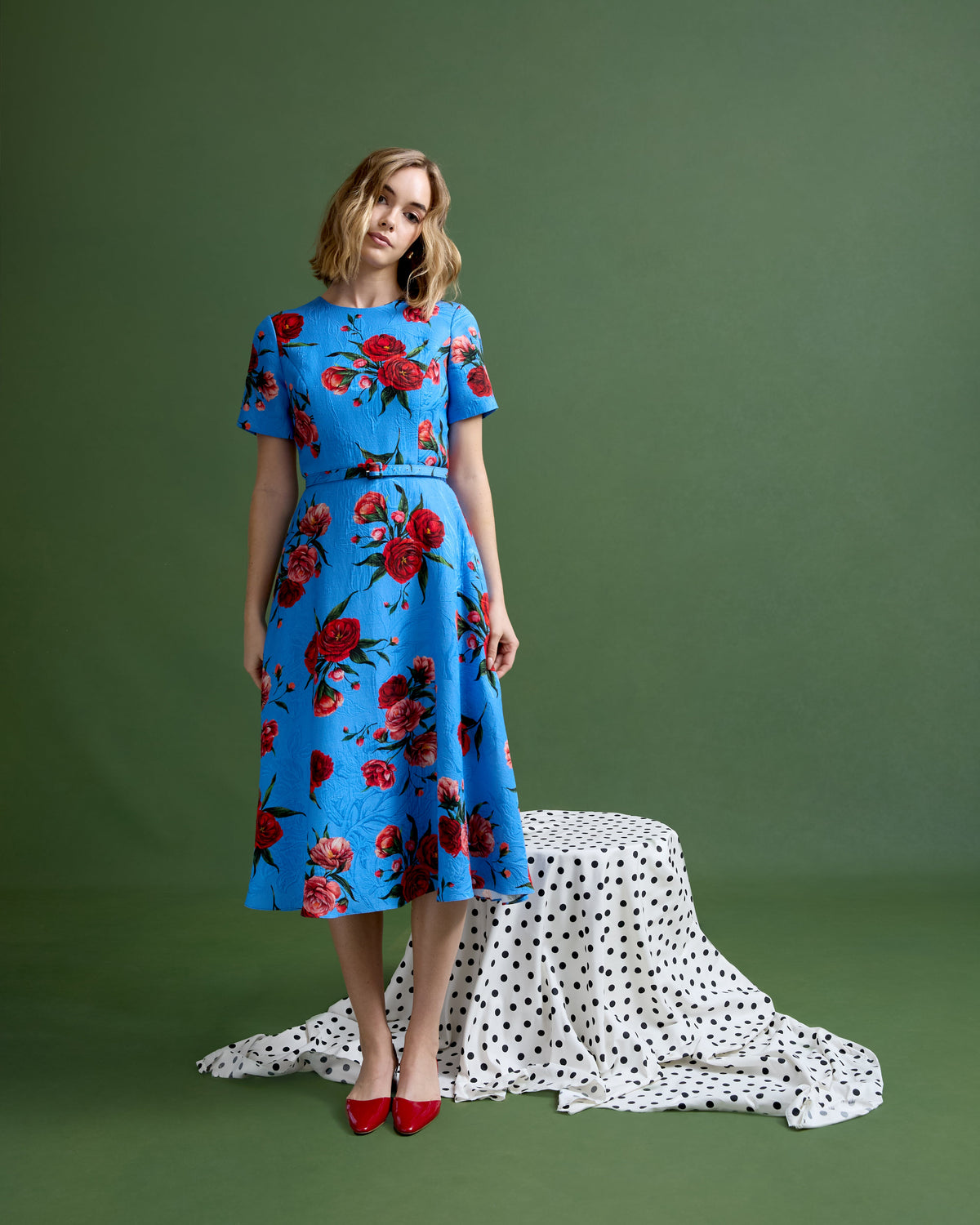 Front view of Harper dress in Chelsea Rose brocade on model, blue brocade with red roses, short sleeves and midi skirt, green backdrop