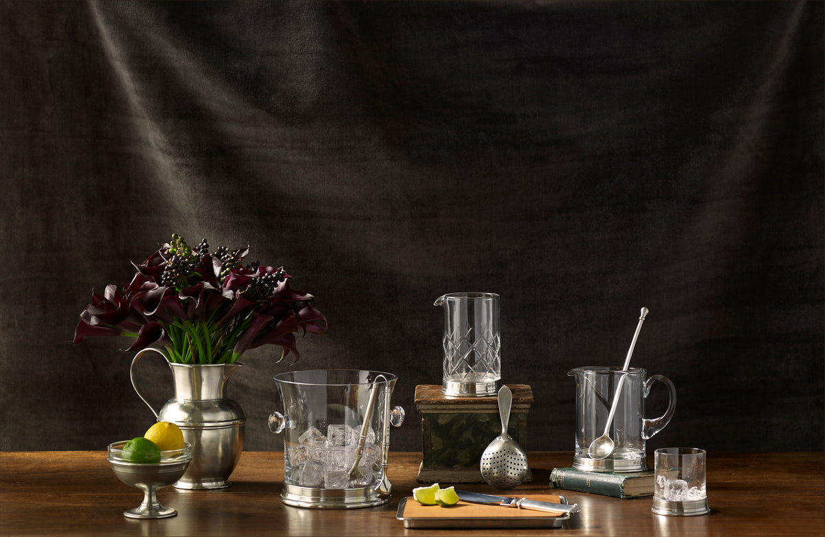 Front view of Branch Bar crystal pitcher and pewter stirrer highlighting pewter accents on wooden table with dark cloth backdrop