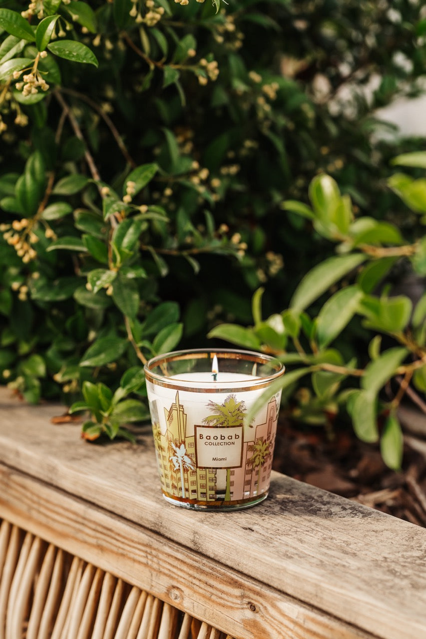 Three-quarter view of Baobab Miami candle in gold Art Deco glass with lit wick on a wooden ledge and green foliage