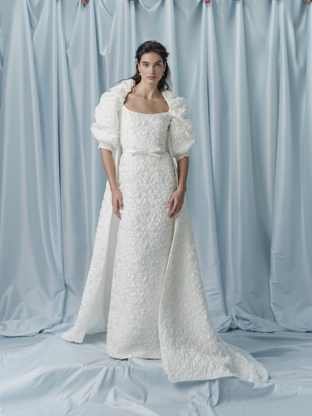 Front view of Adrianna Puff Sleeve Coat showcasing floral jacquard sleeves and fully lined train against blue drapery