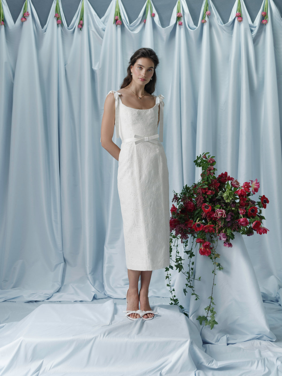 Front view of Viola ivory floral embroidered corset dress showcasing bow belt on model against blue draped backdrop