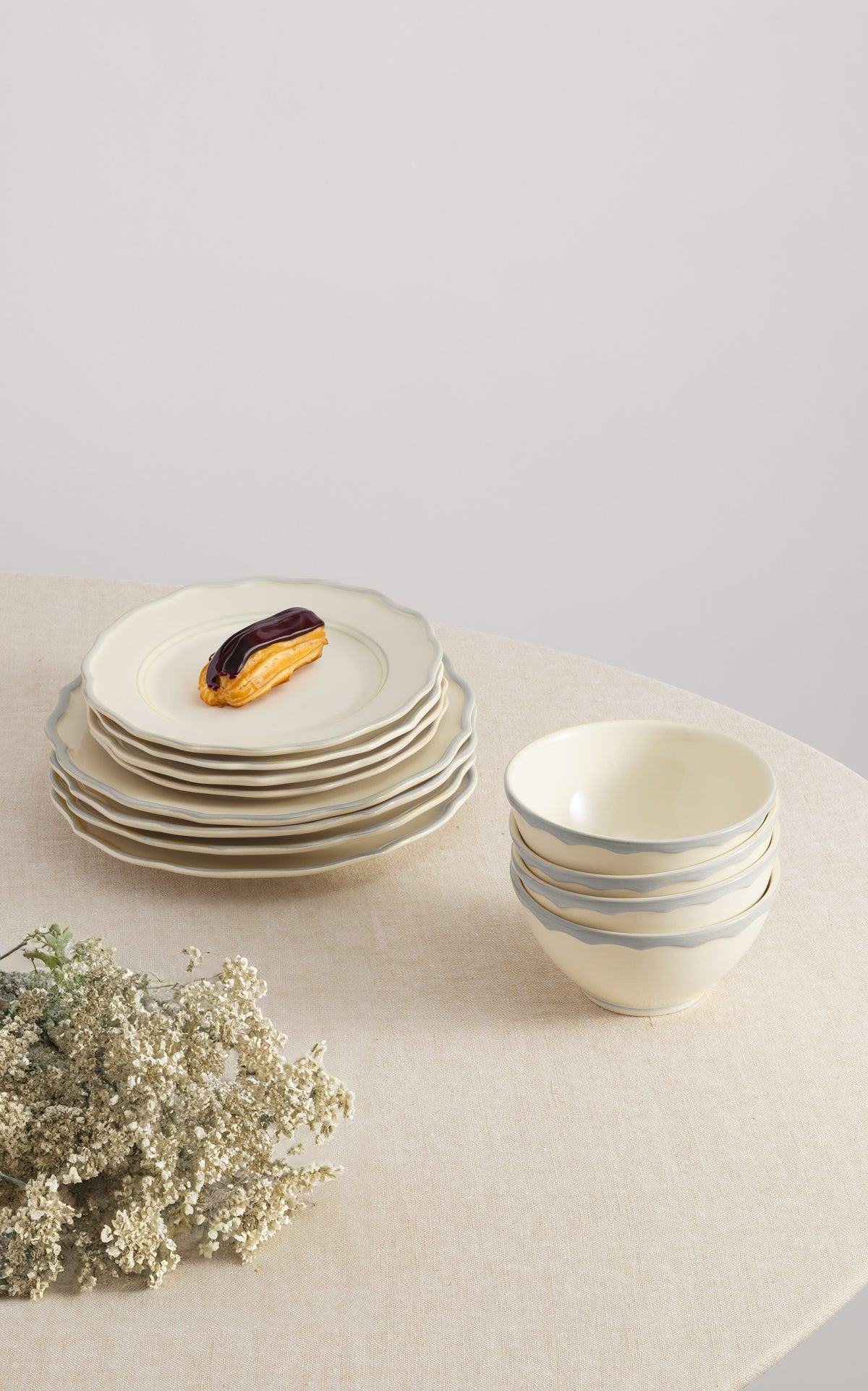 Angled front view of Sylvia 12-piece dinnerware set showing stacked scalloped cream plates and cereal bowls with gray hand-painted rim on a beige tablecloth