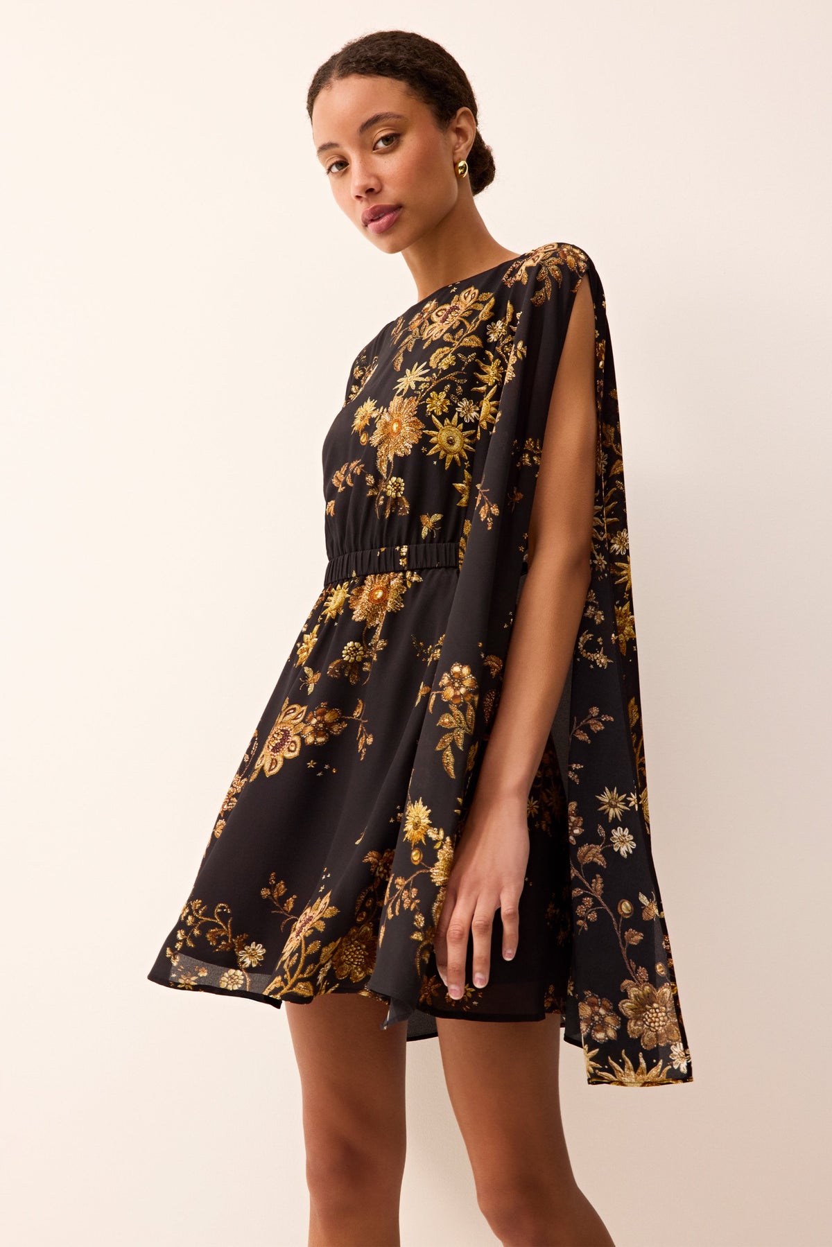 Short black dress with dramatic cape sleeves, cinched waistline, and intricate gold floral print all over.